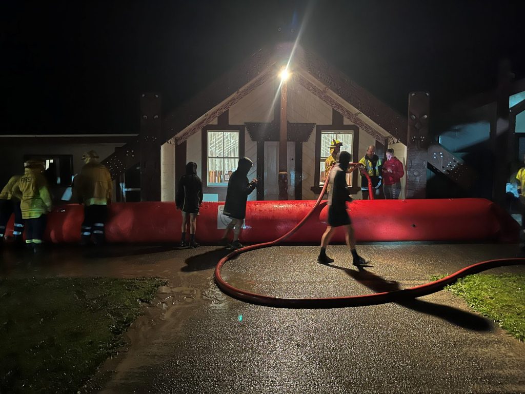 Community members and firefighters working around a red inflatable flood barrier outside Kauwhata Marae at night