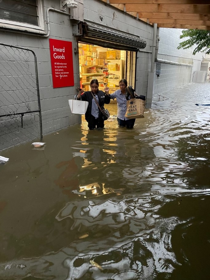 Two supermarket staff walking through deep floodwater outside the inward goods entrance at Fresh Collective Alberton